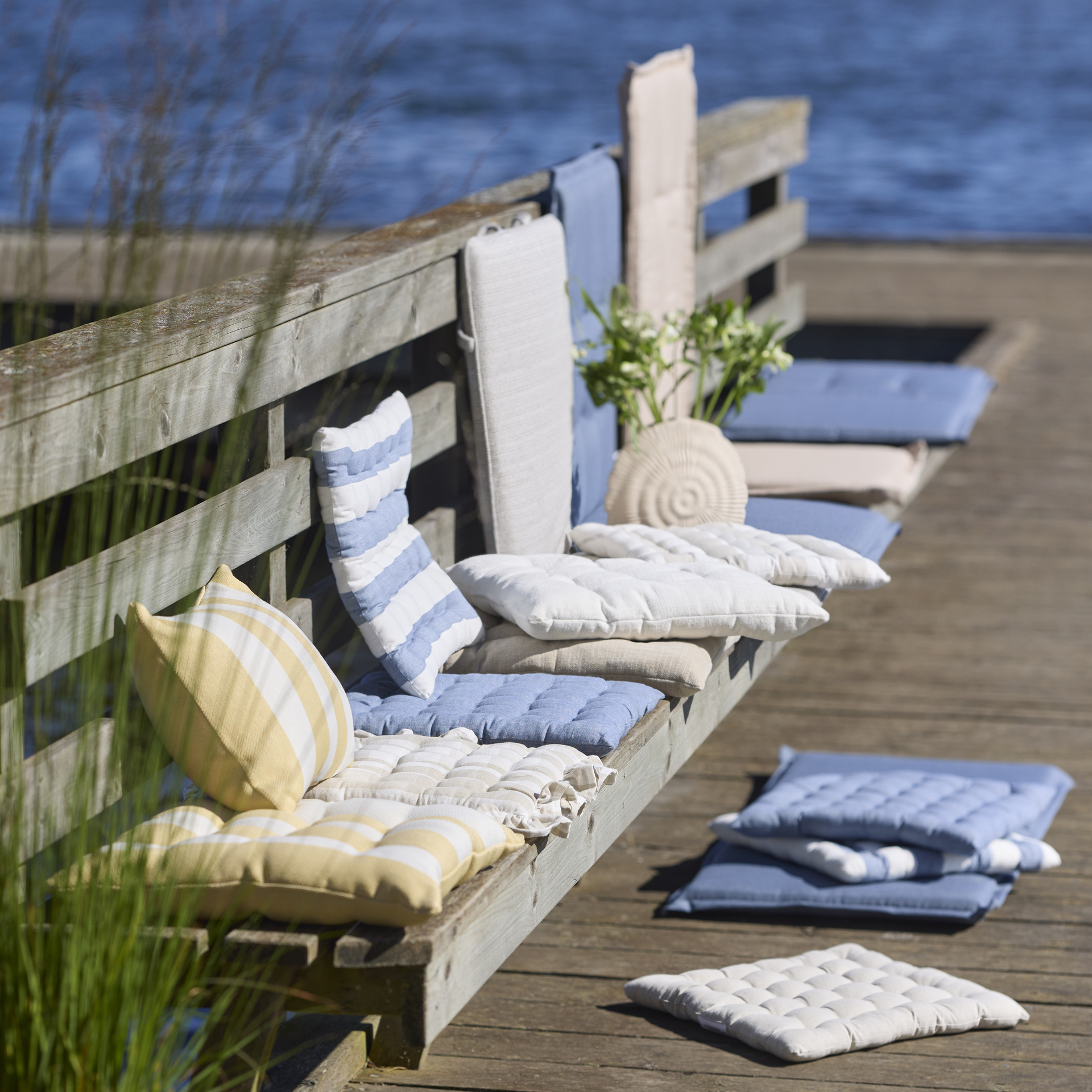 dynor och kuddar i olika mönster och färger ligger på en bänk på en brygga vid vattnet 