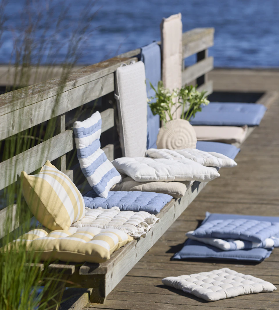 dynor och kuddar i olika mönster och färger ligger på en bänk på en brygga vid vattnet