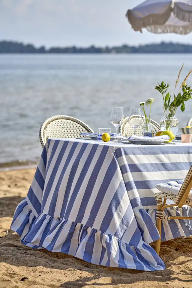 ett bord står dukat vid strandkanten med en blårandig duk, porslin och en vas med gröna kvistar i