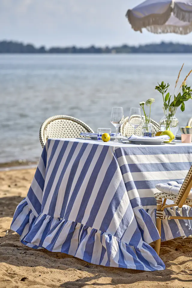 ett bord står dukat vid strandkanten med en blårandig duk, porslin och en vas med gröna kvistar i