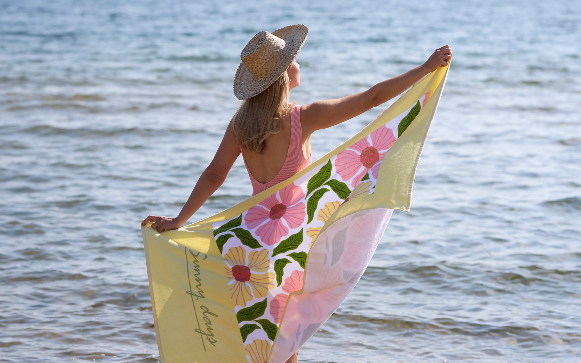 En blond tjej står i en rosa baddräkt på stranden och håller upp en gul och rosa handduk bakom sig och tittar ut mot vattnet