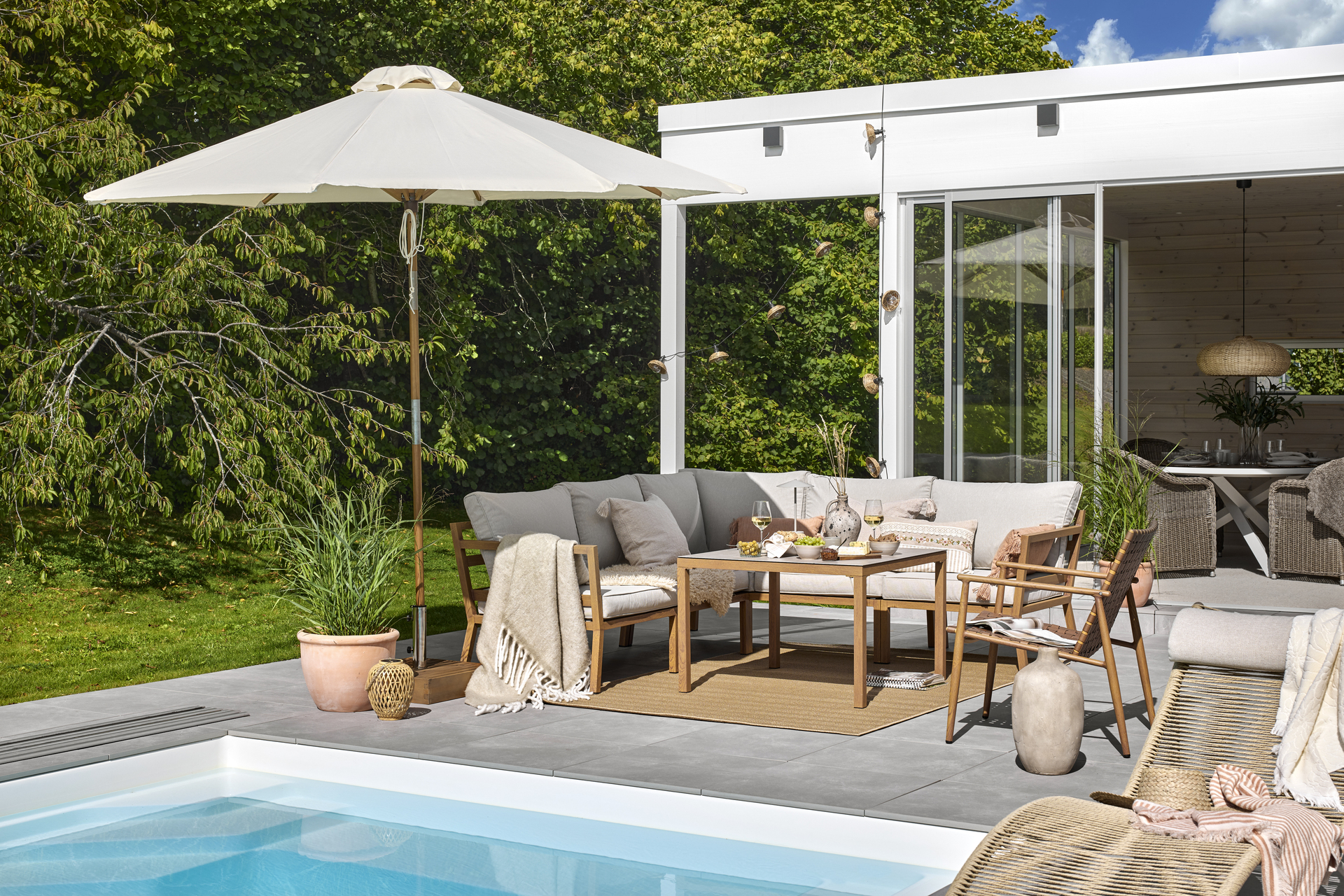 Uteplats med loungemöbler under ett vitt parasoll ståendes framför en pool med blått vatten. 
