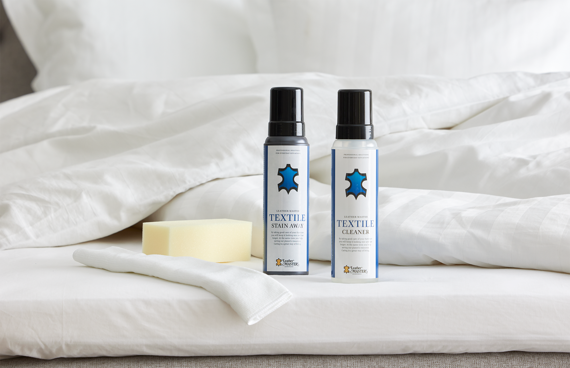 Två flaskor möbelvård med blå logotyper står uppställda på en säng intill en tvättsvamp och fibertrasa. 