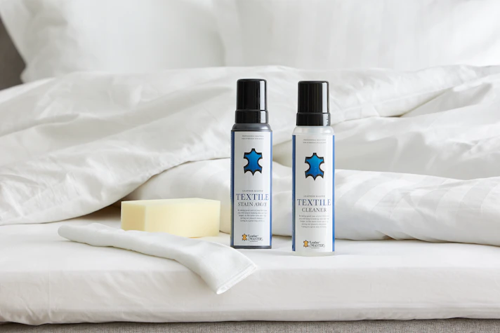 Två flaskor möbelvård med blå logotyper står uppställda på en säng intill en tvättsvamp och fibertrasa.