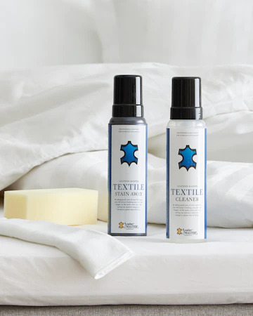 Två flaskor möbelvård med blå logotyper står uppställda på en säng intill en tvättsvamp och fibertrasa.