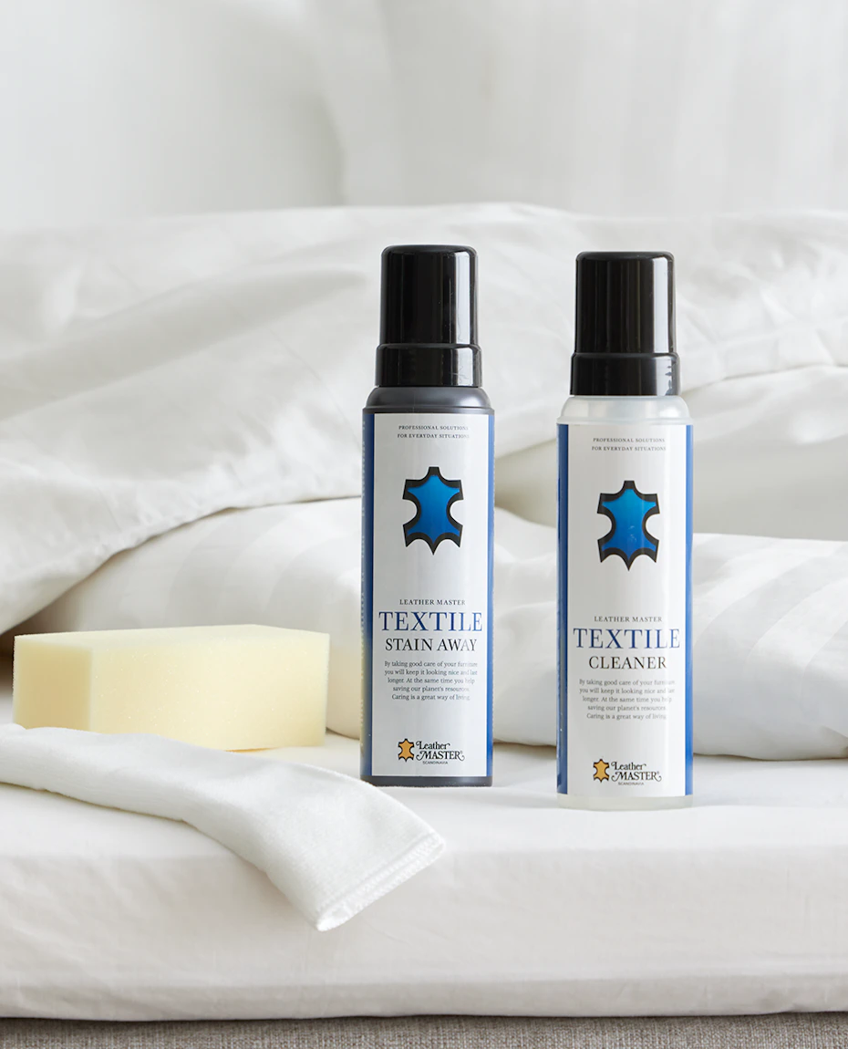 Två flaskor möbelvård med blå logotyper står uppställda på en säng intill en tvättsvamp och fibertrasa.