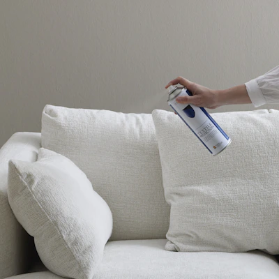 en hand sticker fram och håller i en sprayflaska och sprayar mot en vit soffa