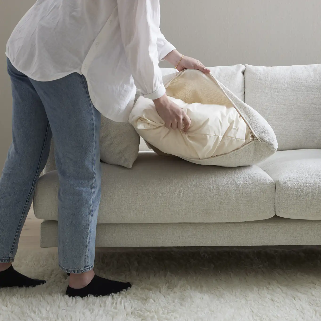 En person i ljusa jeans och vit skjorta tar av klädseln på en av kuddarna till en vit soffa