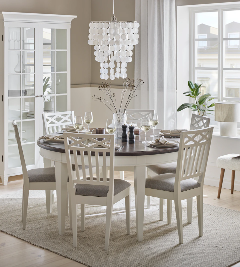 Ett runt vitt bord med bordsskiva i mörkt trä och sex matchande stolar samt ett vitt vitrinskåp står i ett ljust matrum.