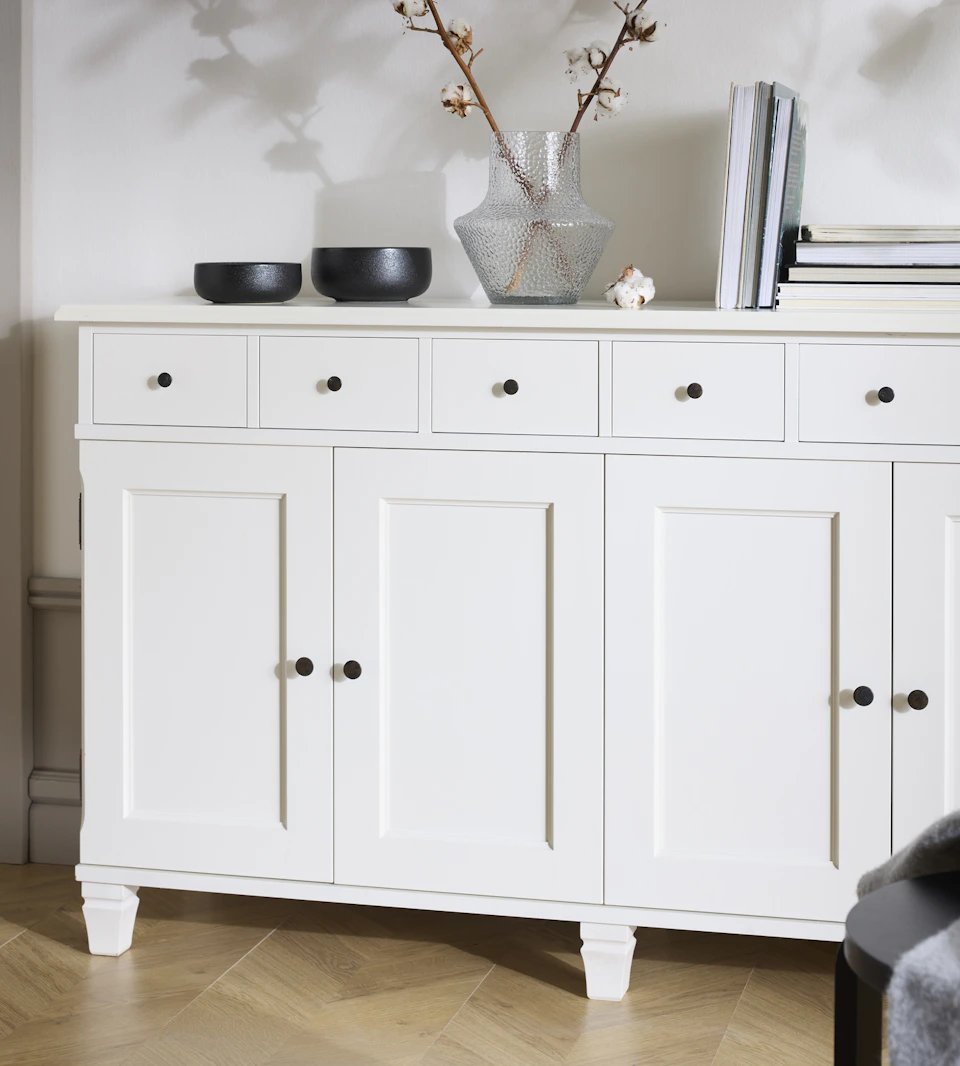 En vit skänk med två skåp och flera lådor står framför en vit vägg.