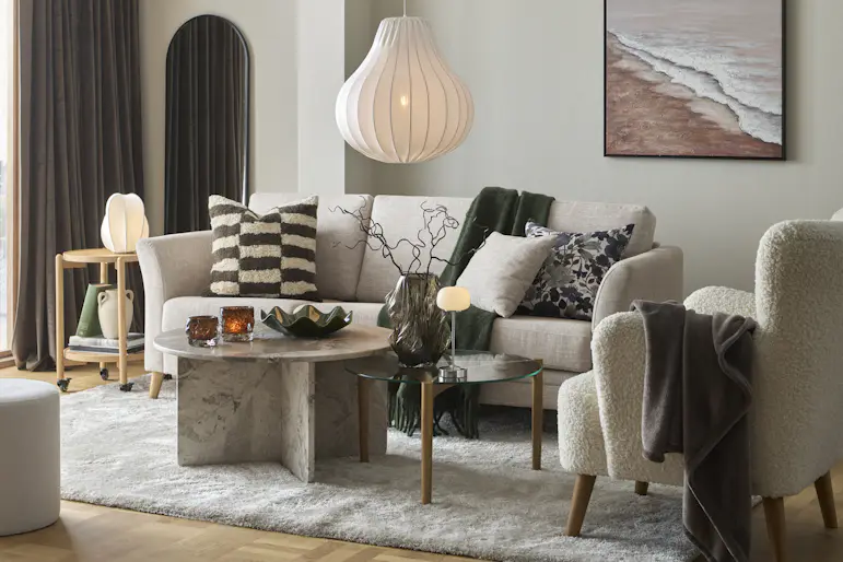 Ett vardagsrum där det står en beige soffa, fåtölj, soffbord i marmor och en stor vit matta