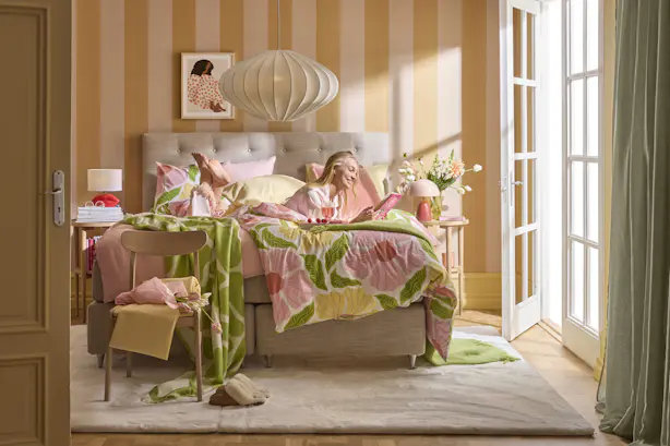 en blond tjej ligger i en beige säng i ett färgglatt sovrum och läser en bok