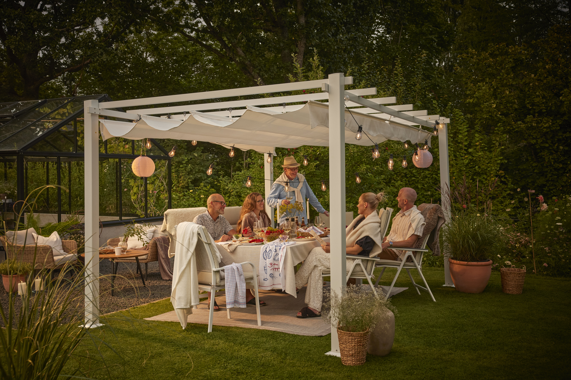 en stort gäng människor sitter och står runt ett bord under en vit pergola i en trädgård