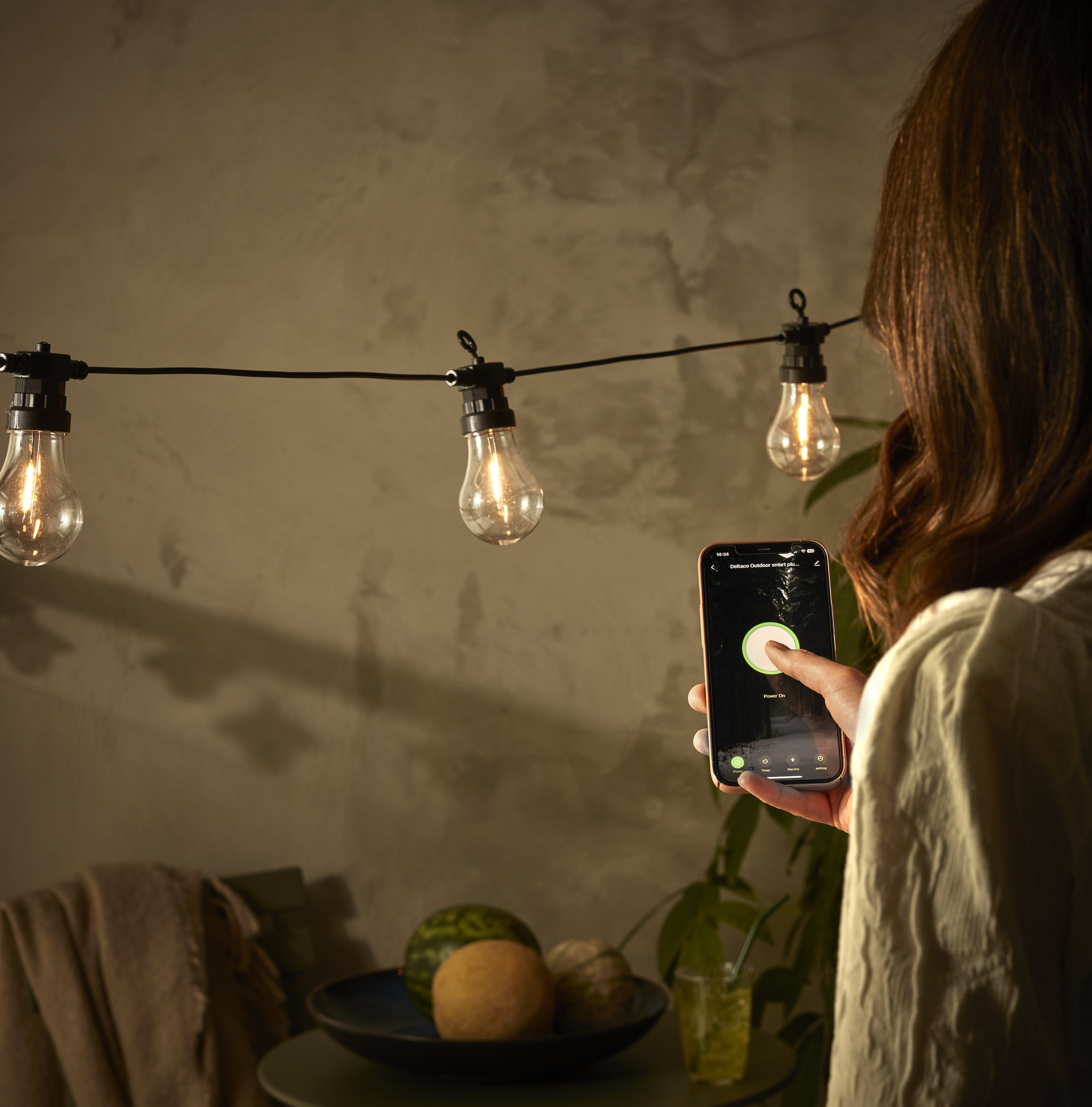 en ljusslinga hänger ute framför en stenvägg och en hand håller upp en telefon och styr belysningen