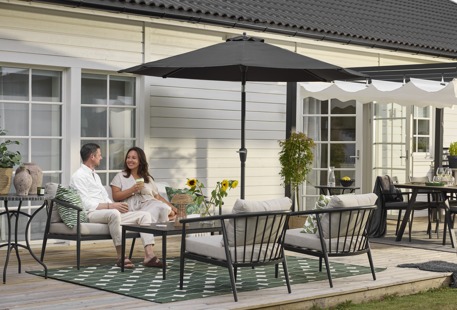 På en altan i en modern, svart loungegrupp med beiga dynor sitter en man och en kvinna och dricker drinkar