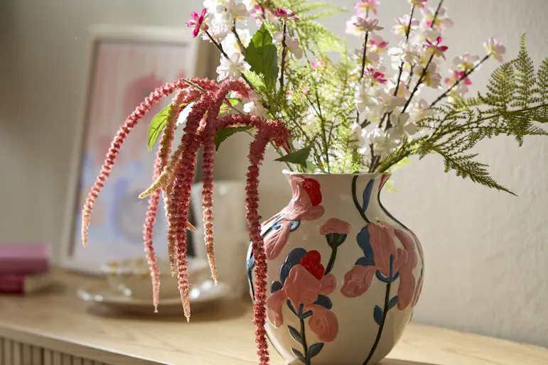 En konstgjord bukett står i en vit vas målad med rosa blommor.