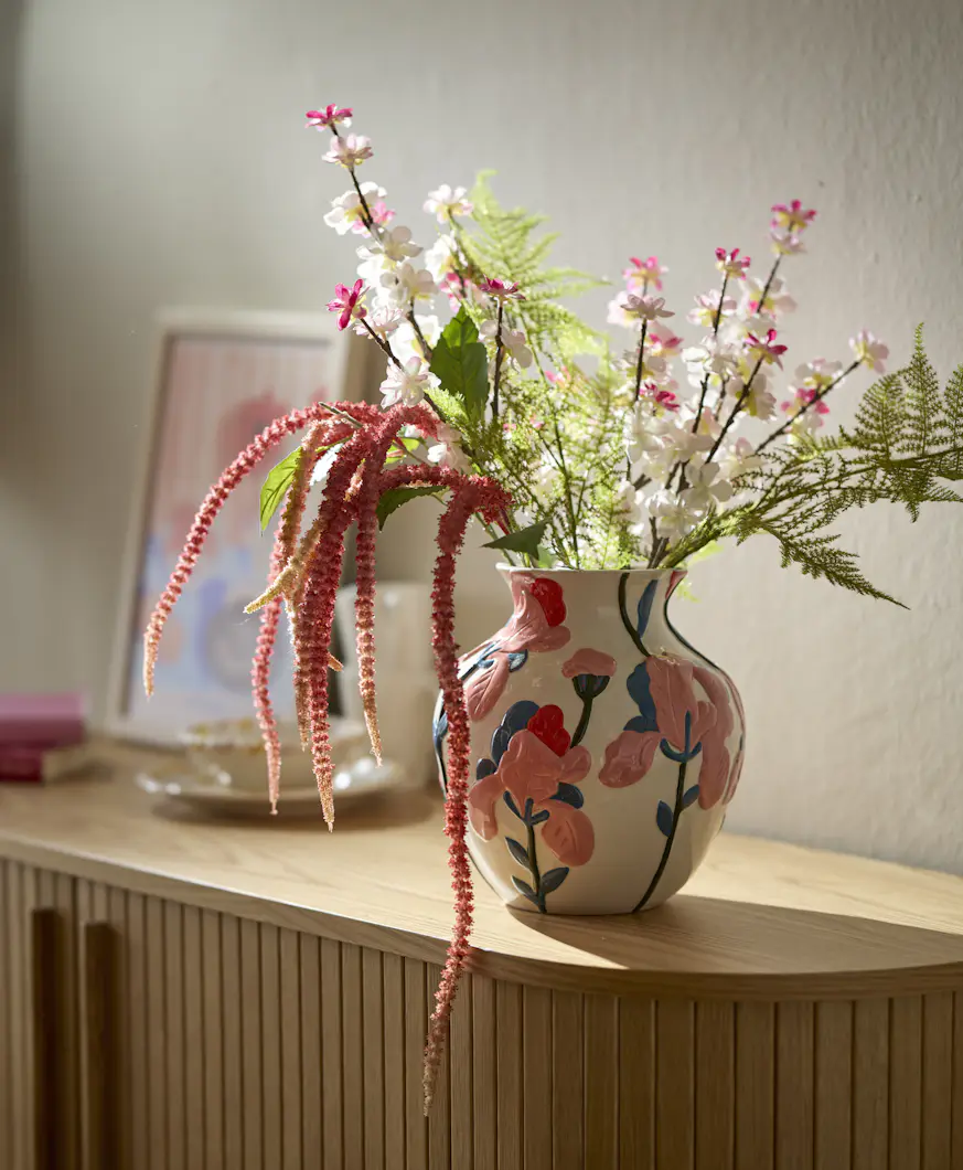 En konstgjord bukett står i en vit vas målad med rosa blommor.