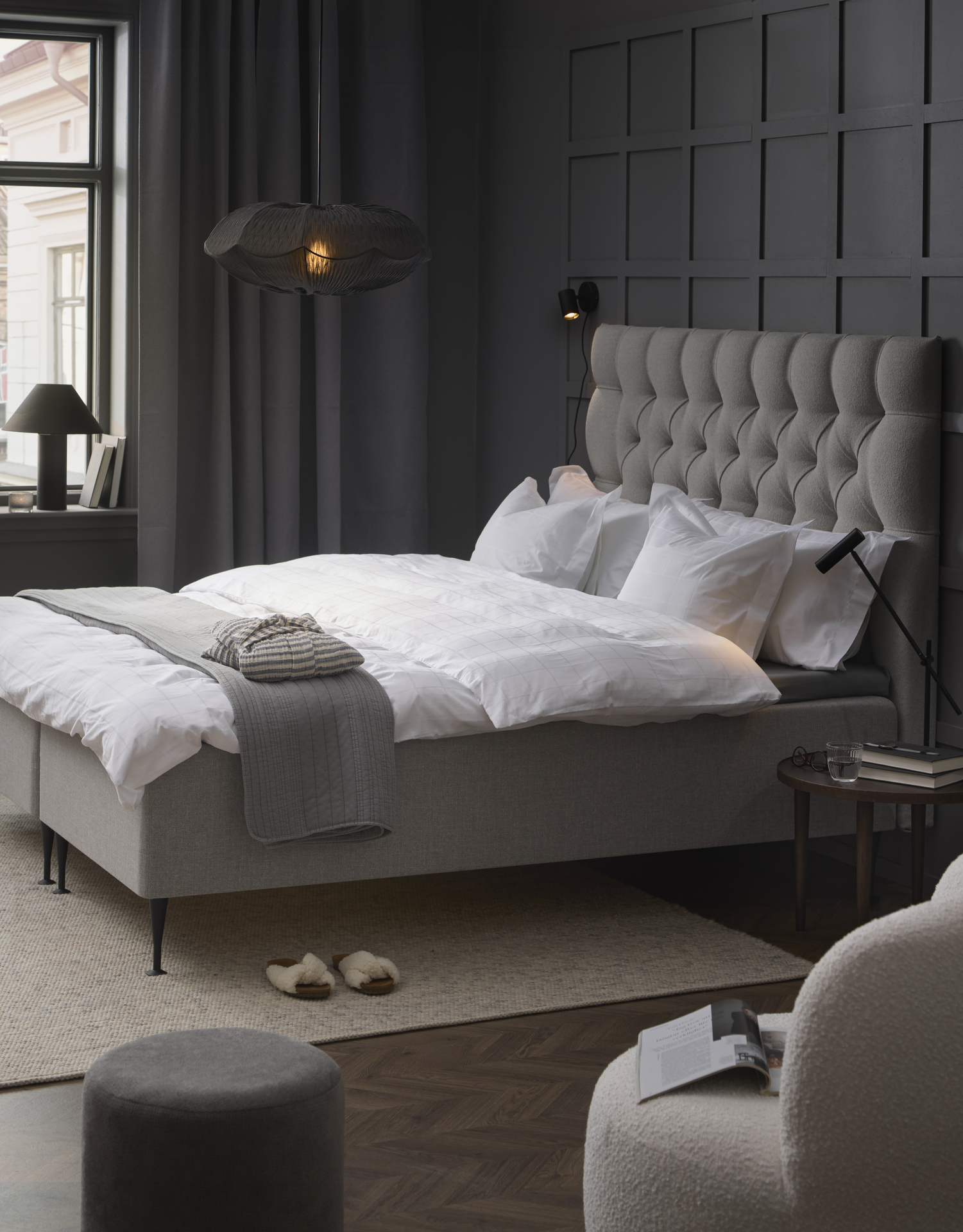 Stilrent och stramt sovrum med grå väggar och mitt i rummet en kontinentalsäng i grått tyg med pikerad sänggavel. 