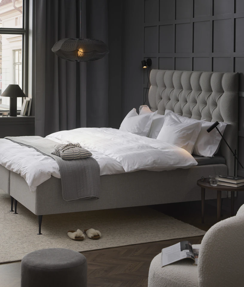 Stilrent och stramt sovrum med grå väggar och mitt i rummet en kontinentalsäng i grått tyg med pikerad sänggavel.