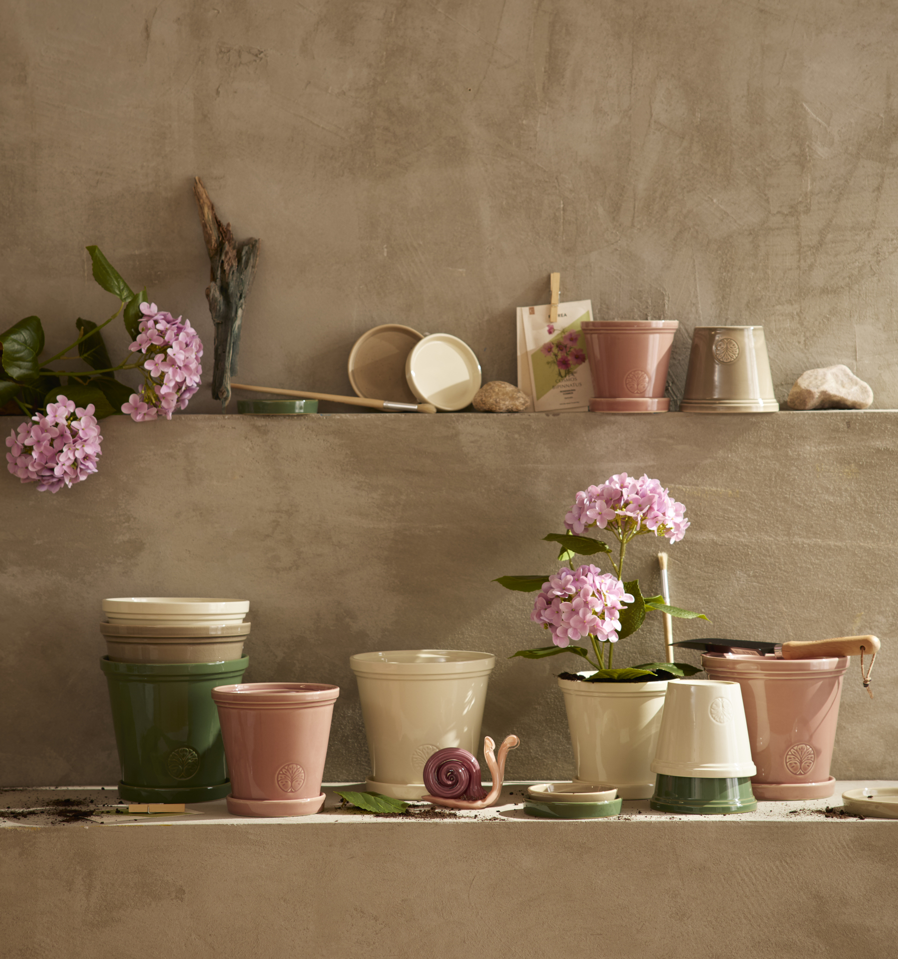 Många cremevita, gammelrosa och gröna krukor i olika storlekar står uppställda framför en putsvägg. I en av krukorna finns en konstgjord rosa hortensia.
