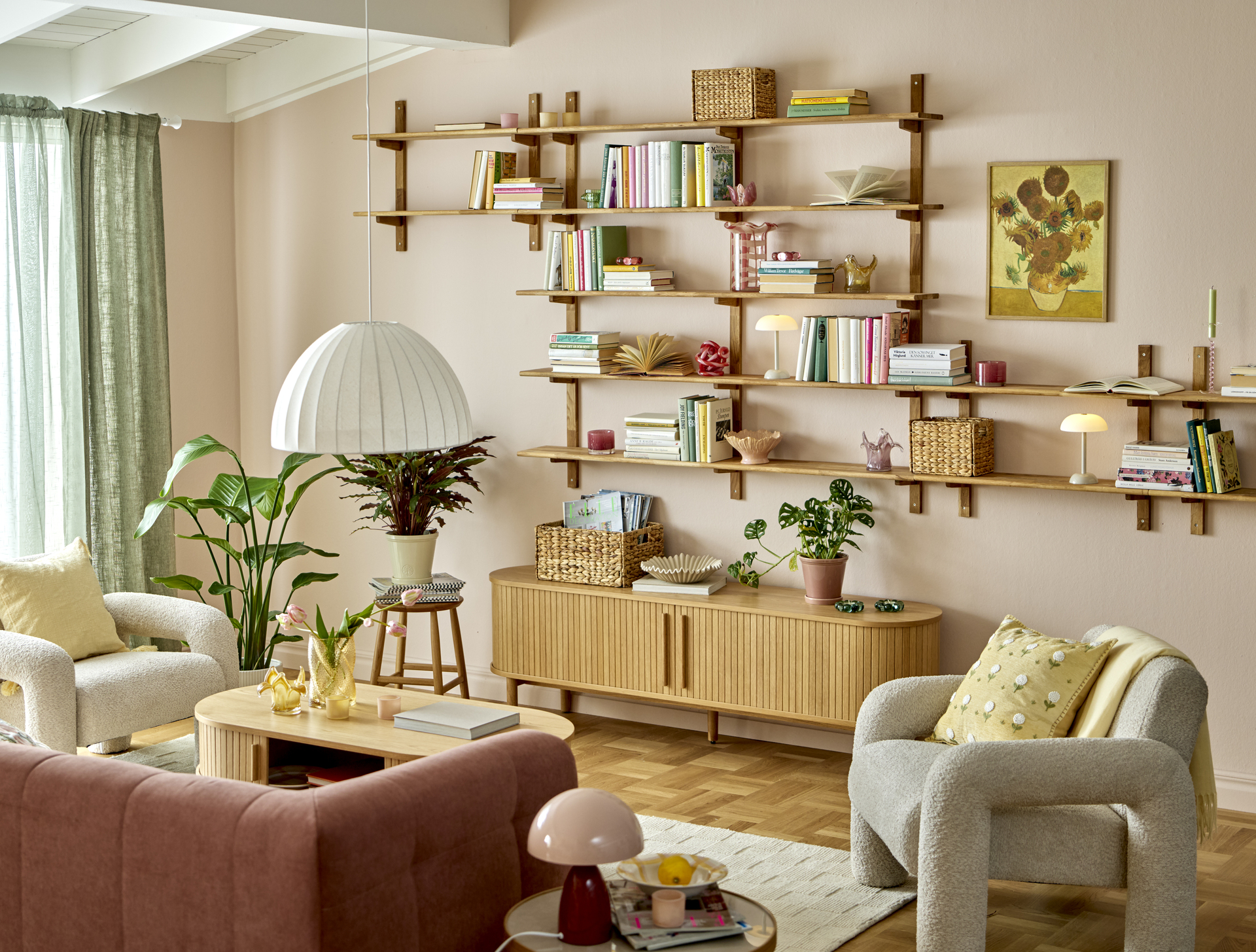 Ett ljust vardagsrum inrett med rosa soffa, beiga fåtöljer, ett soffbord med en vas tulpaner, en stor krukväxt på golvet, en mindre krukväxt på en pall, en tv-bänk med magasin och en krukväxt samt en stor vägghylla.