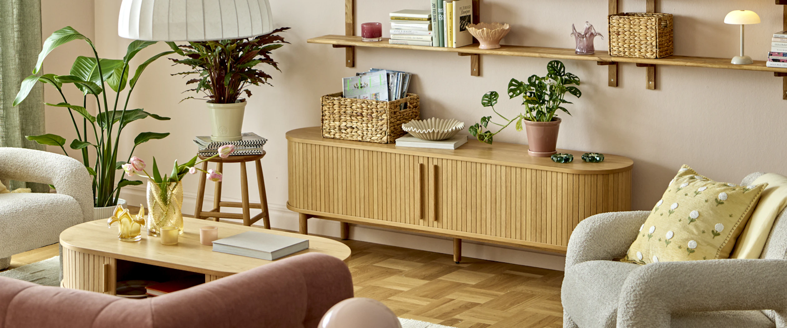 Ett ljust vardagsrum inrett med rosa soffa, beiga fåtöljer, ett soffbord med en vas tulpaner, en stor krukväxt på golvet, en mindre krukväxt på en pall, en tv-bänk med magasin och en krukväxt samt en stor vägghylla.
