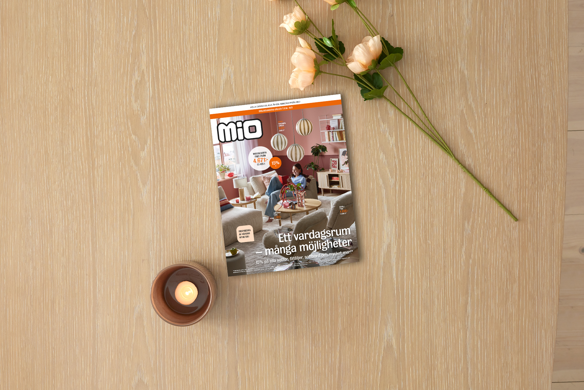 På ett ljust träbord ligger kampanjbladet för Mios vardagsrumskampanj intill ett ljus och en blomma.