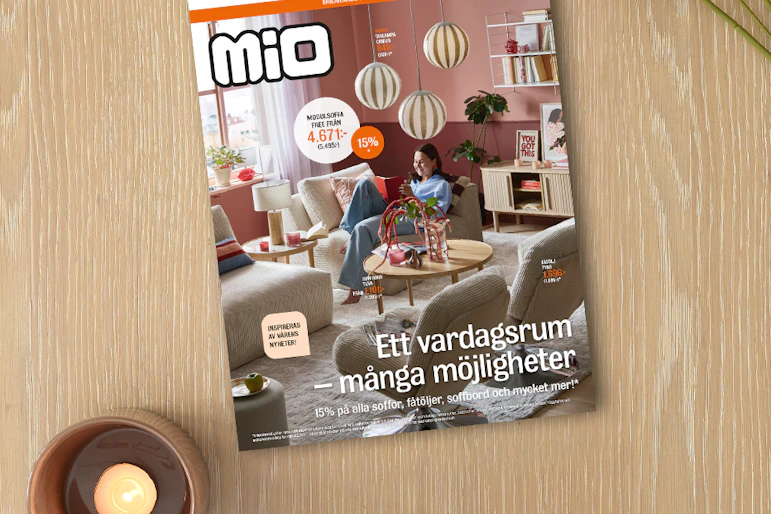 På ett ljust träbord ligger kampanjbladet för Mios vardagsrumskampanj intill ett ljus och en blomma.