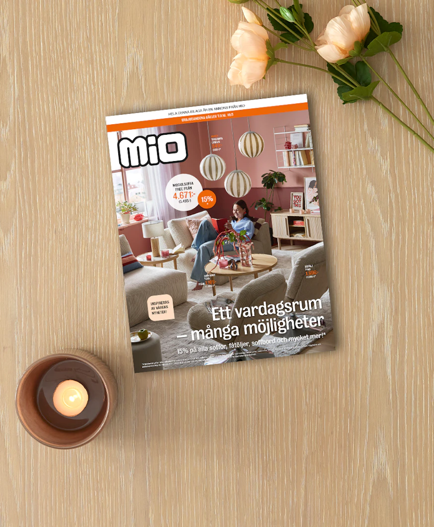 På ett ljust träbord ligger kampanjbladet för Mios vardagsrumskampanj intill ett ljus och en blomma.