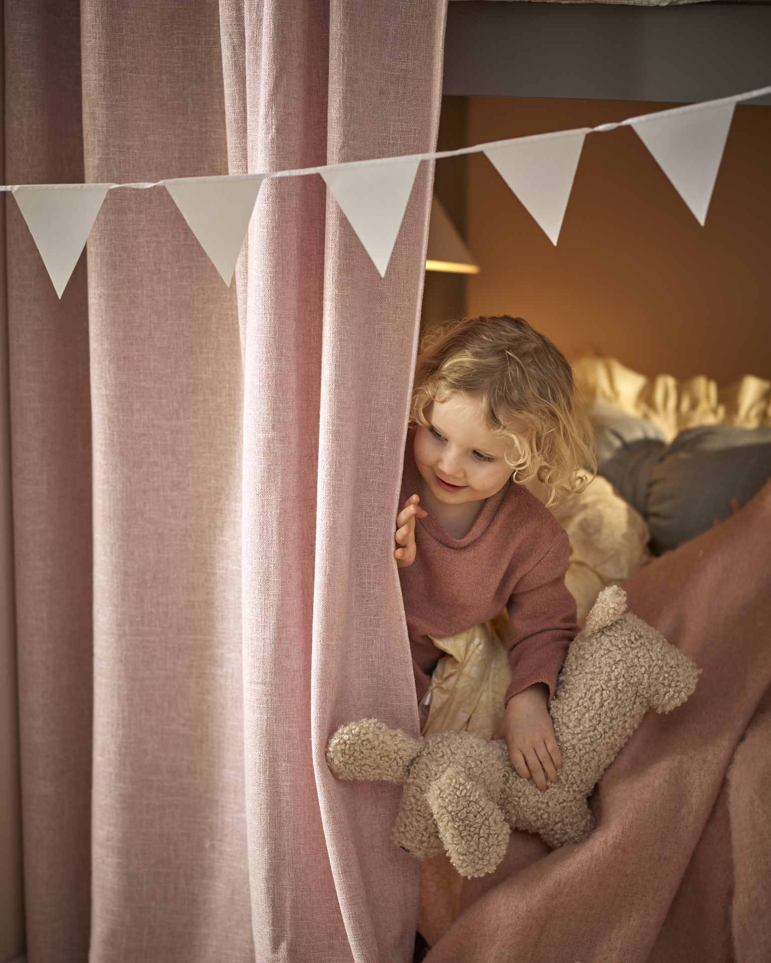 Ett barn sitter i en säng och kikar fram bakom en rosa gardin