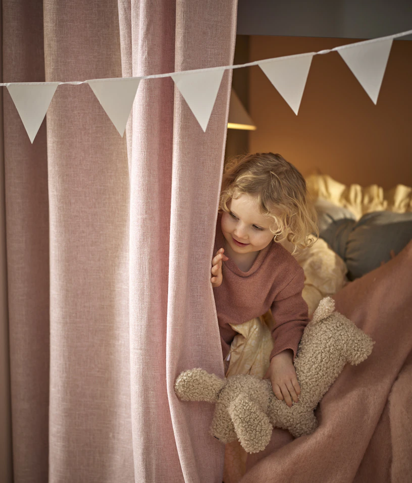 Ett barn sitter i en säng och kikar fram bakom en rosa gardin