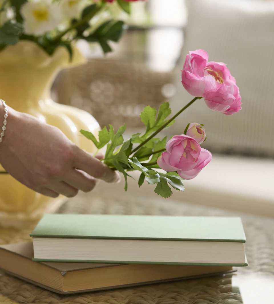 En hand håller i en konstjord rosa ranunkel. I bakgrunden syns en gul vas.