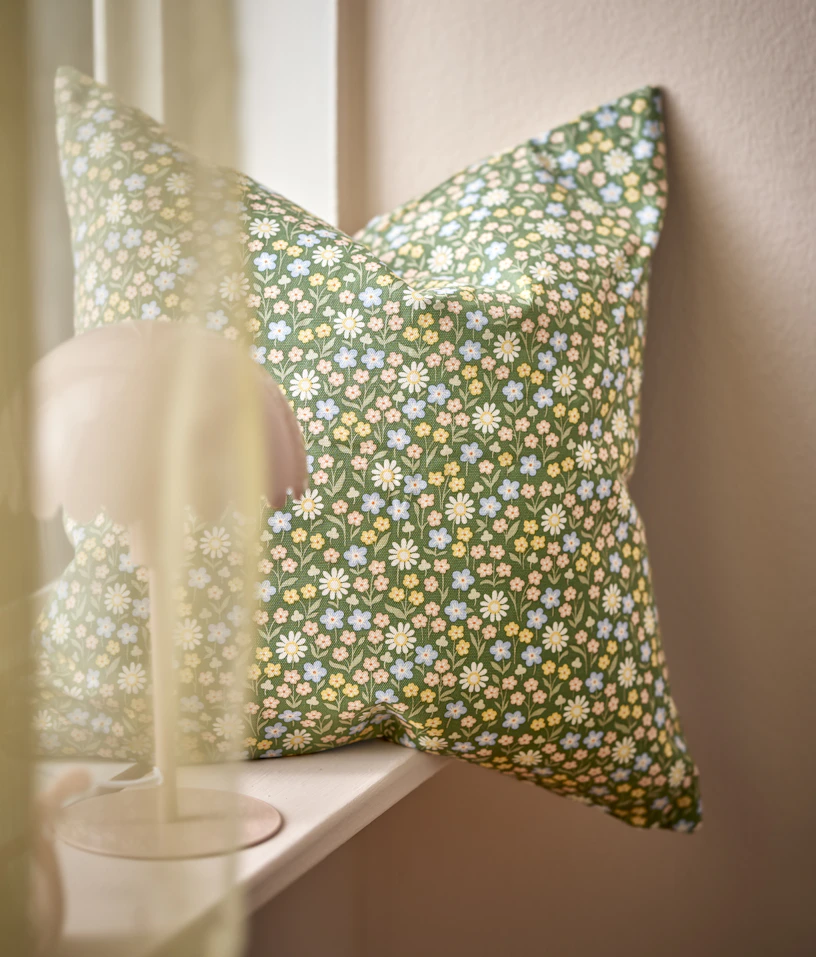 En grön kudde med blommönster står i ett fönster vid en rosa vägg
