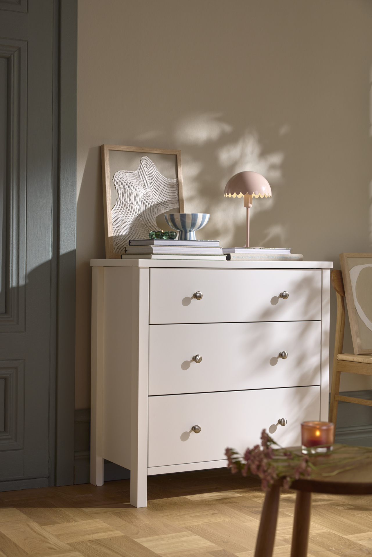 En vit byrå står mot en beige vägg. På byrån finns en lampa, tavla och en hög med tidningar.