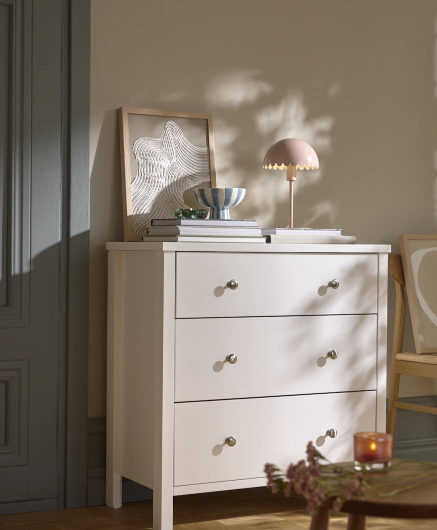 En vit byrå står mot en beige vägg. På byrån finns en lampa, tavla och en hög med tidningar.