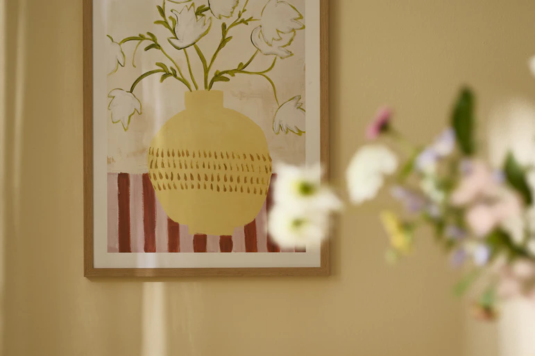 en tavlka med motiv av en vas med blommor hänger på en gul vägg