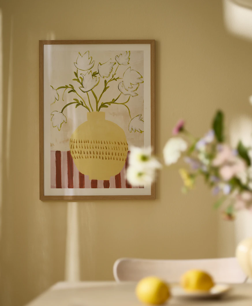 en tavlka med motiv av en vas med blommor hänger på en gul vägg