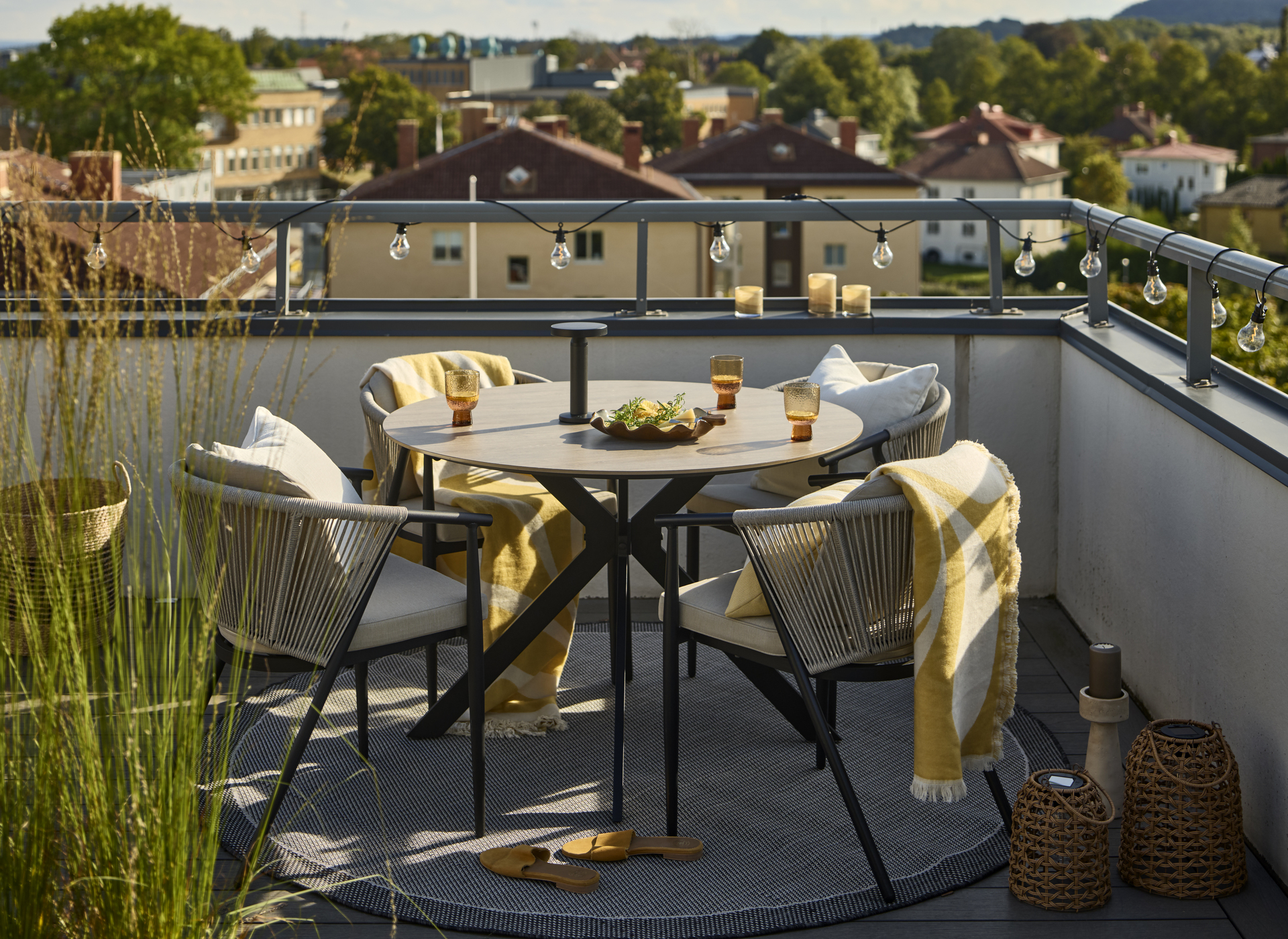 På en balkong står ett runt bord med beige bordskiva och fyra matchande karmstolar med gula plädar. En ljusslinga är virad runt balkongräcket.