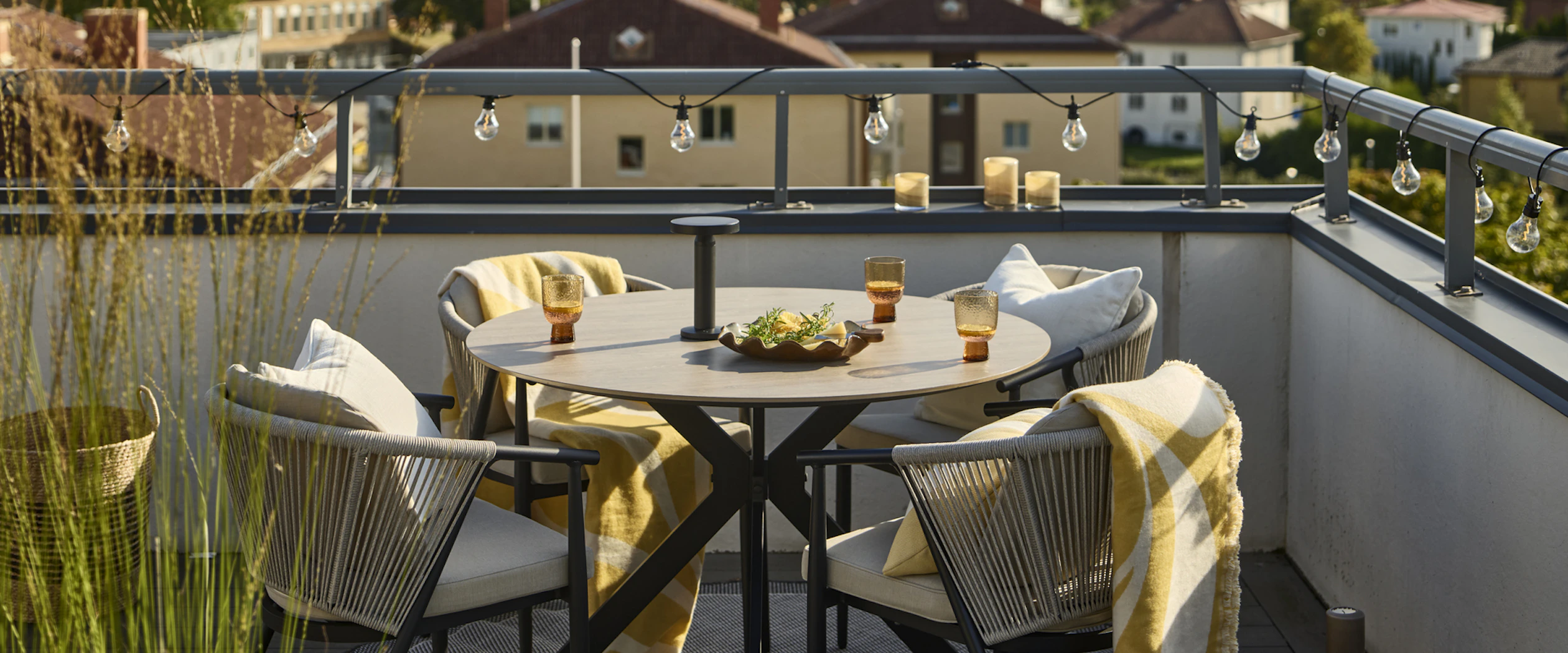 På en balkong står ett runt bord med beige bordskiva och fyra matchande karmstolar med gula plädar. En ljusslinga är virad runt balkongräcket.