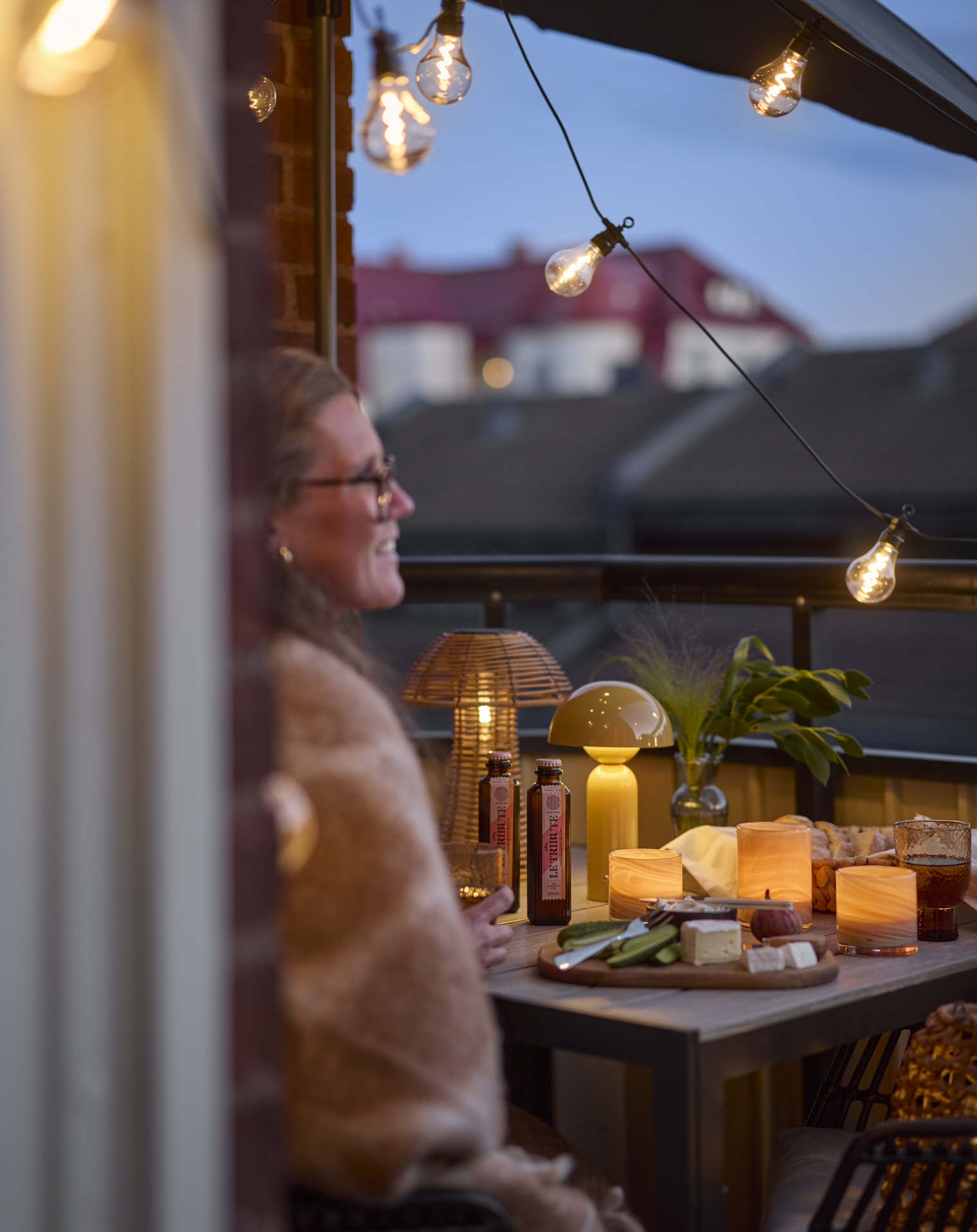 En kvinna sitter vid ett bord på en balkong i kvällsljus. En ljusslinga och två portabla bordslampor lyser.