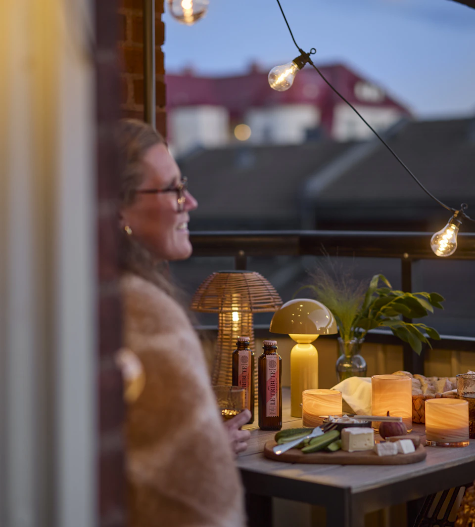 En kvinna sitter vid ett bord på en balkong i kvällsljus. En ljusslinga och två portabla bordslampor lyser.
