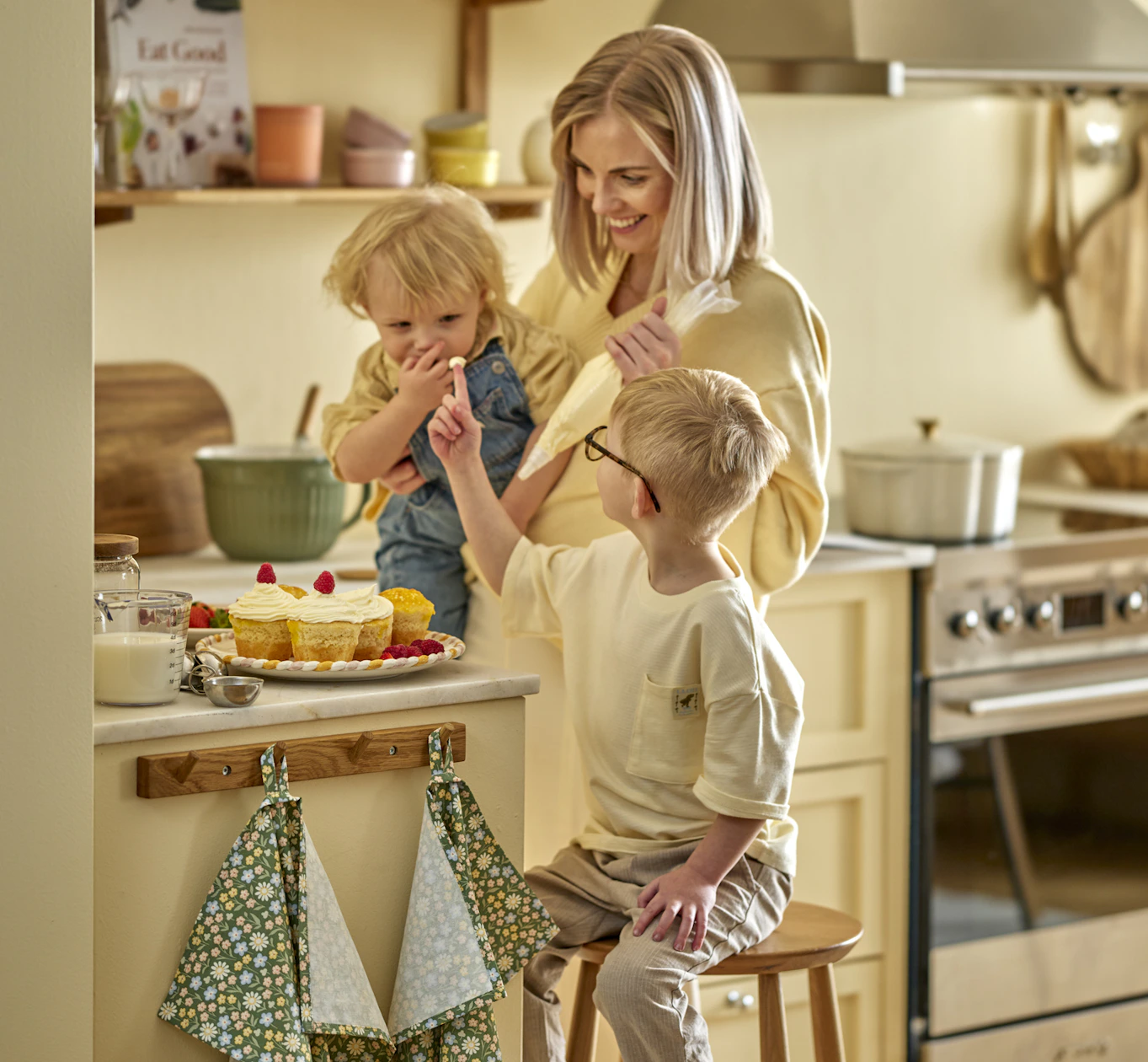 en blond tjej står i ett kök med två små barn och bakar en tårta