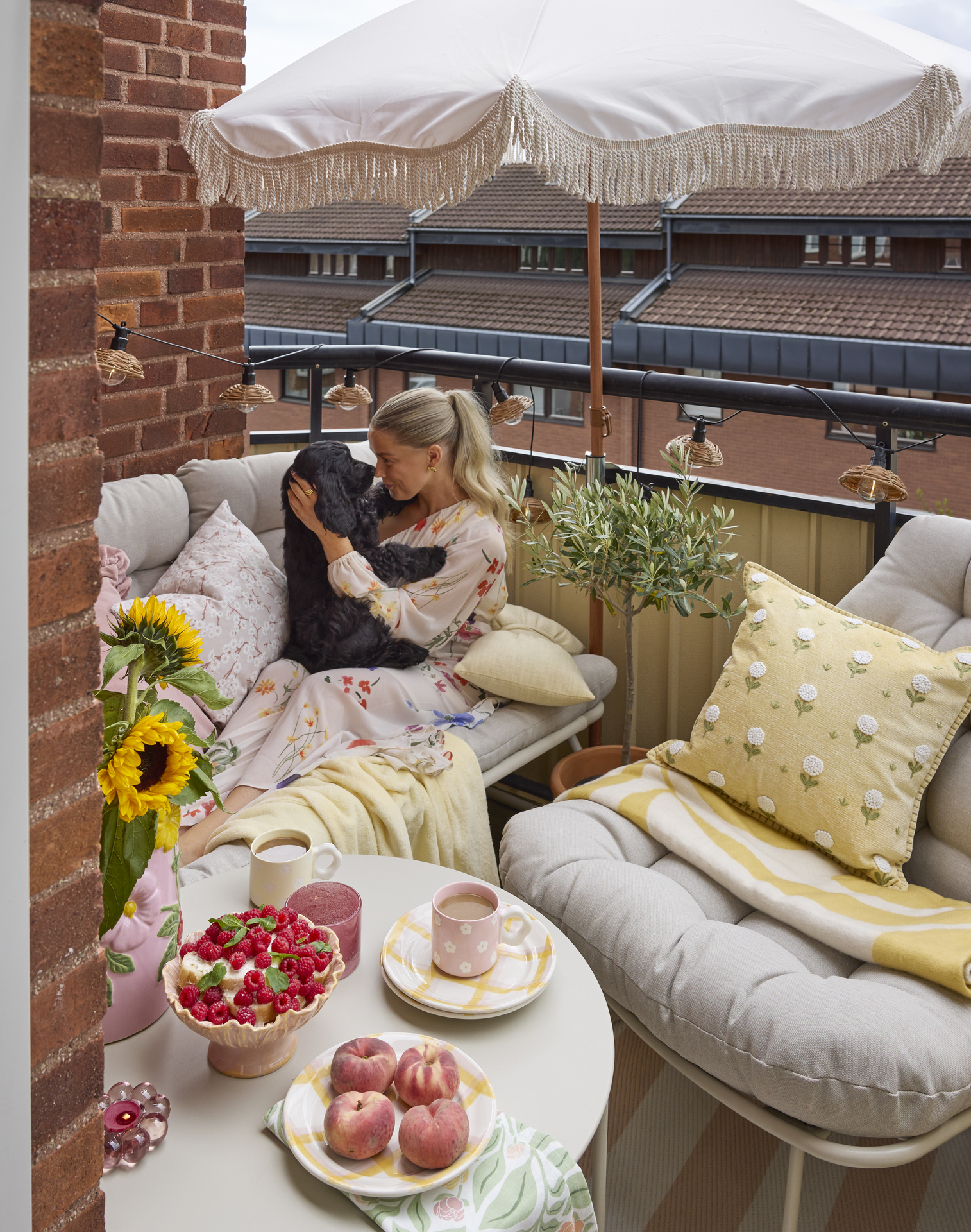 En balkong är möblerad med en beige liten soffa och en matchande fåtölj med gula plädar och kuddar, ett parasoll och en ljusslinga runt balkongräcket. I soffan sitter en tjej och gosar med en svart hund. 