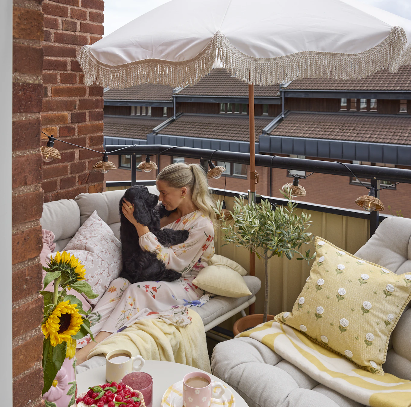 En balkong är möblerad med en beige liten soffa och en matchande fåtölj med gula plädar och kuddar, ett parasoll och en ljusslinga runt balkongräcket. I soffan sitter en tjej och gosar med en svart hund.