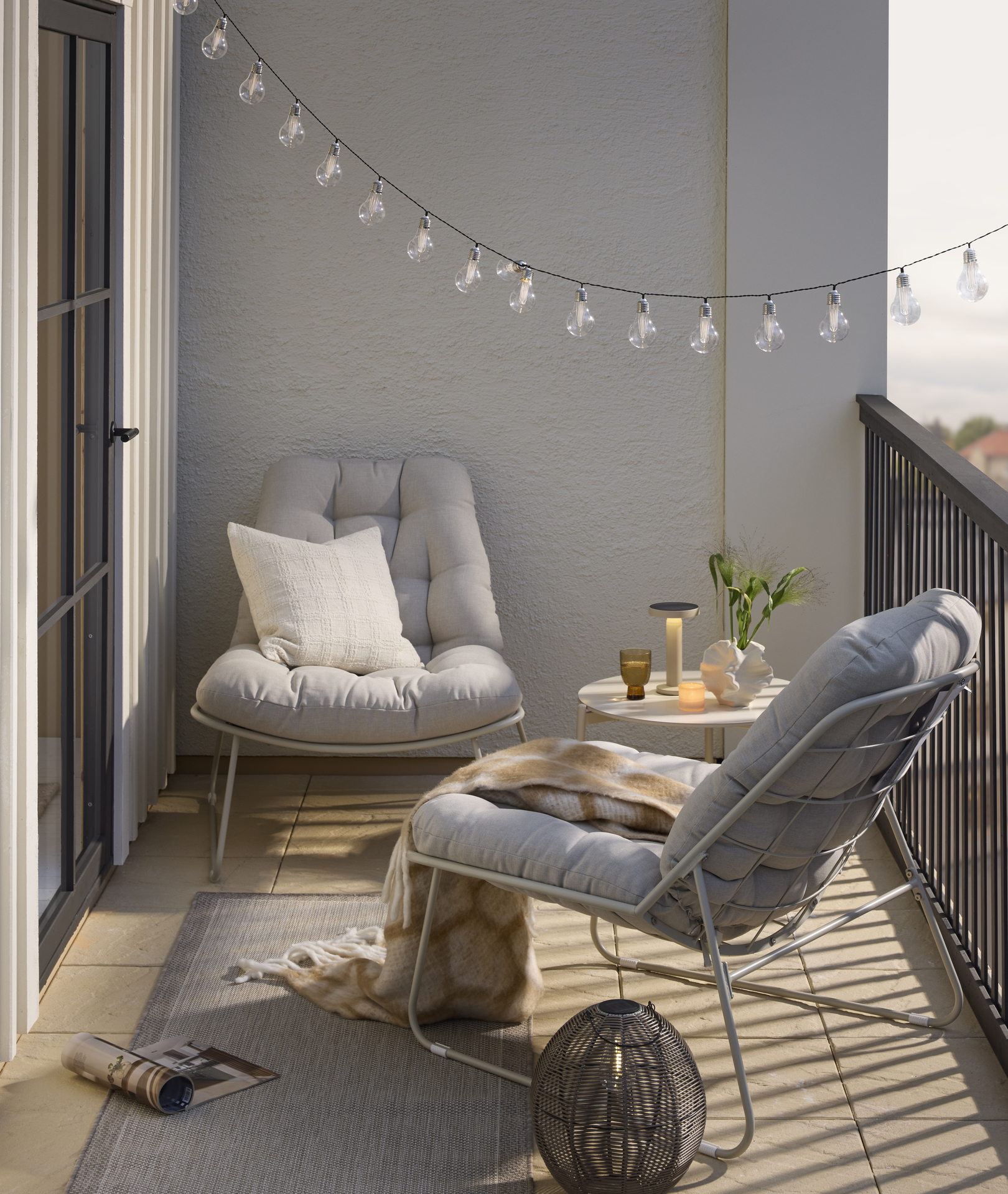 En balkong är möblerad med två beiga fåtöljer, ett runt bord med en portabel lampa och en ljusslinga.
