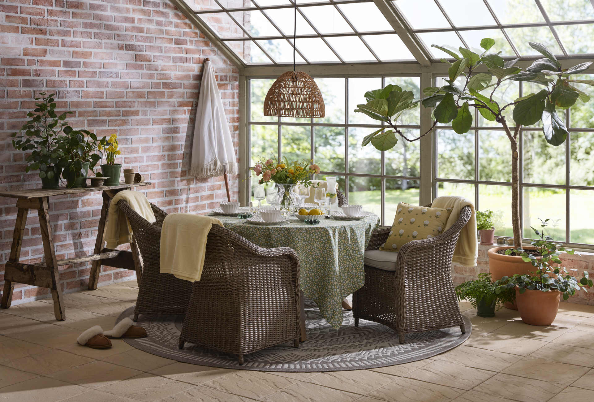 En utesoffgrupp med runt bord och fyra fåtöljer i konstrotting står i ett orangeri fyllt av växter.