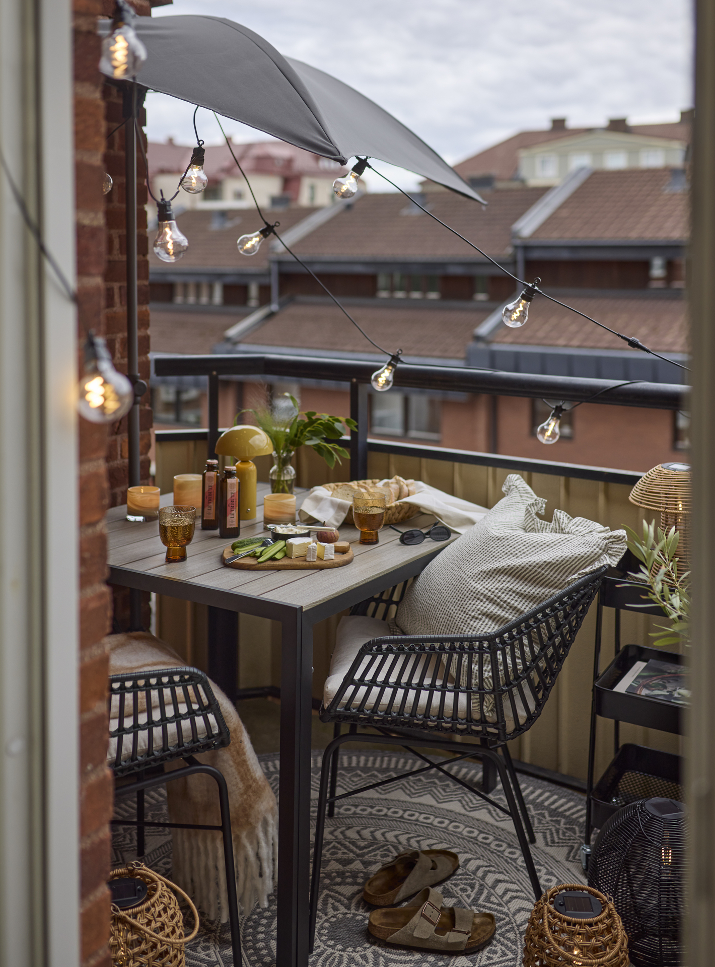 En liten balkong är möblerad med ett kvadratiskt bord och två karmstolar, ett litet parasoll, en ljusslinga och en rullvagn.