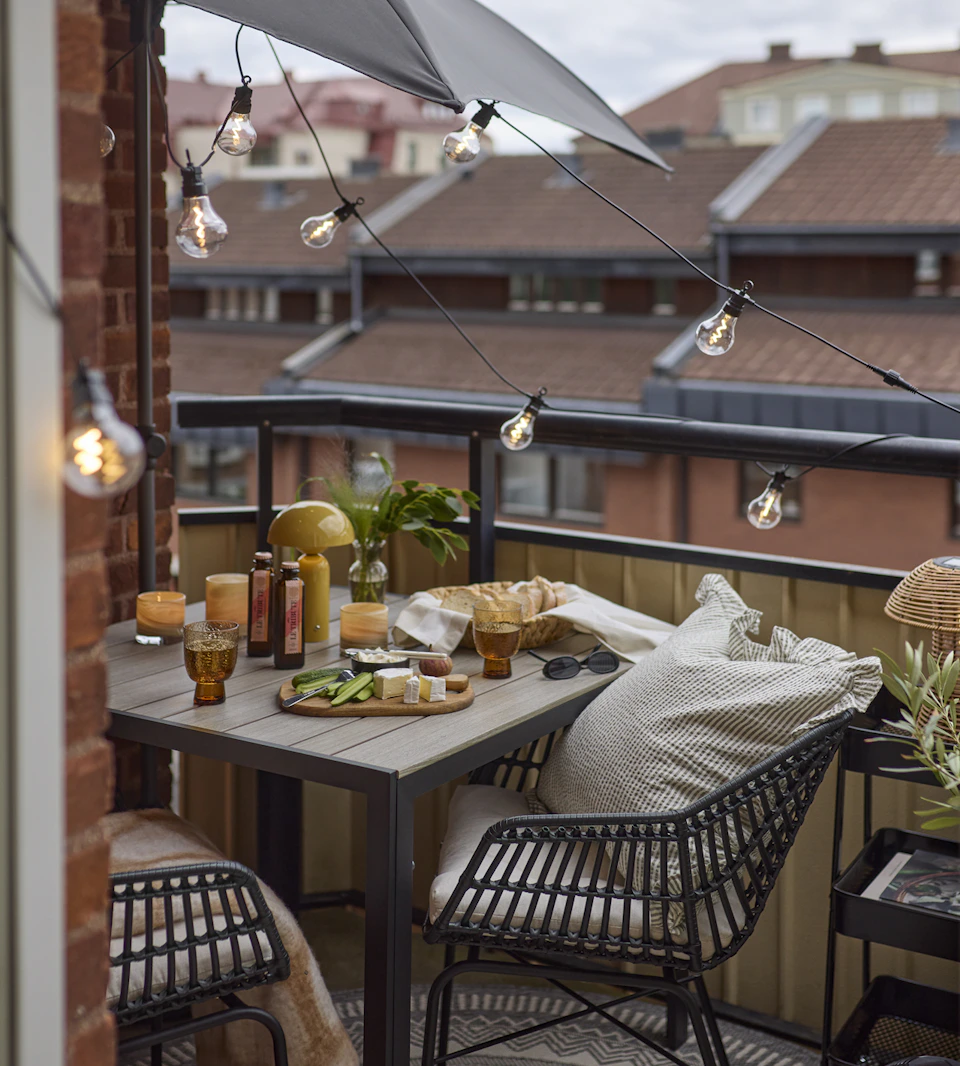 En liten balkong är möblerad med ett kvadratiskt bord och två karmstolar, ett litet parasoll, en ljusslinga och en rullvagn.