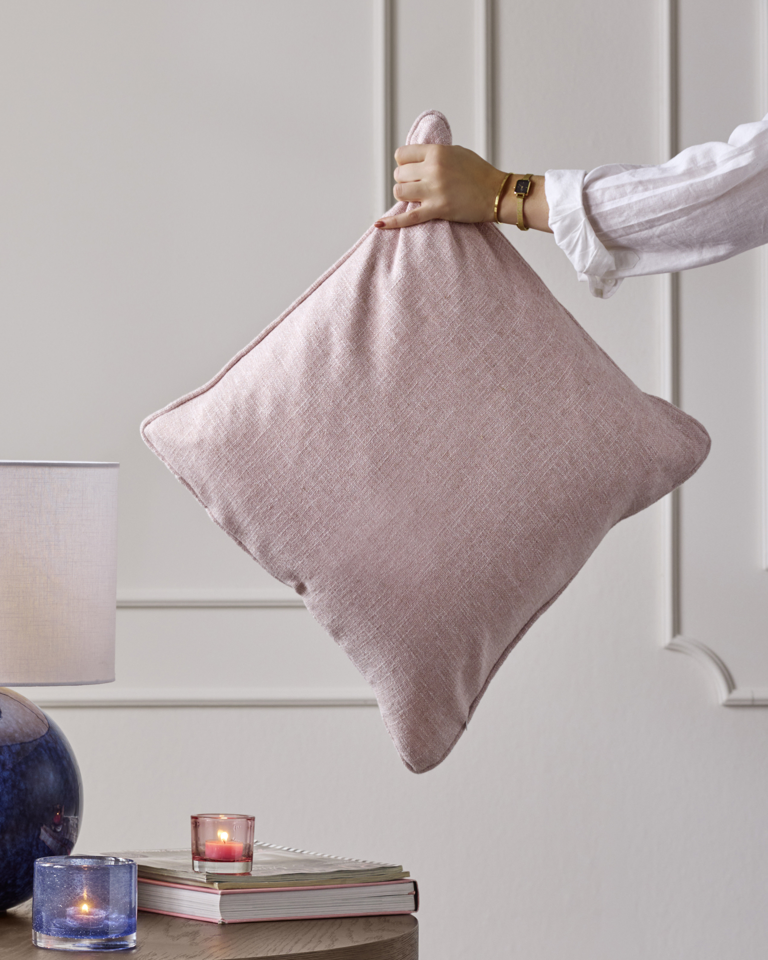 En hand håller i en prydnadskudde med rosa kuddfodral mot en vit vägg.