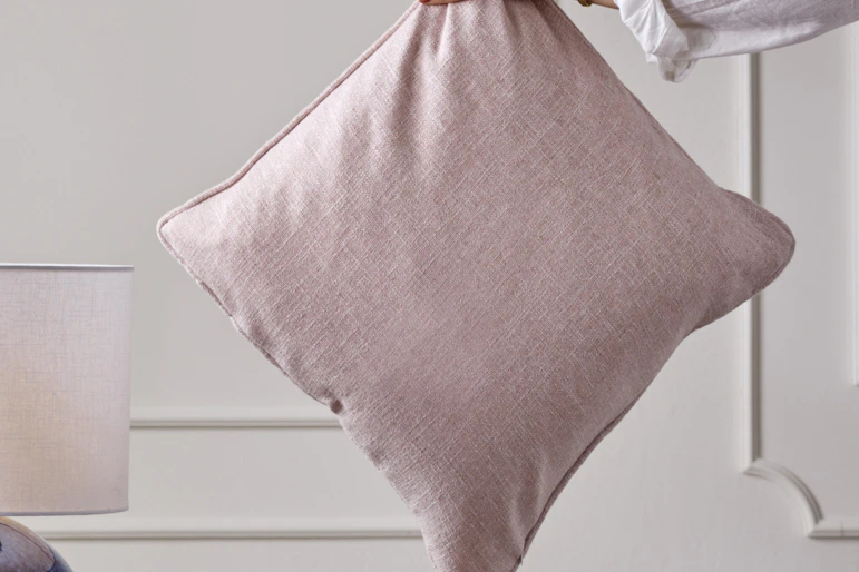 En hand håller i en prydnadskudde med rosa kuddfodral mot en vit vägg.